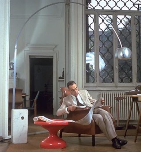 Achille Castiglioni in his studio sitting under an Arco arc lamp on a Sanluca chair next to a Rochcetto table, all by his hand.